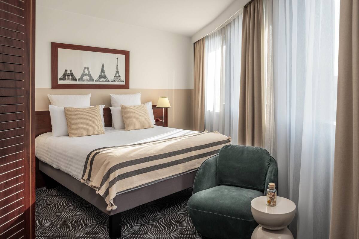 A hotel room with a bed, a green armchair, a small table with a decorative item, and a framed picture of the Eiffel Tower on the wall.