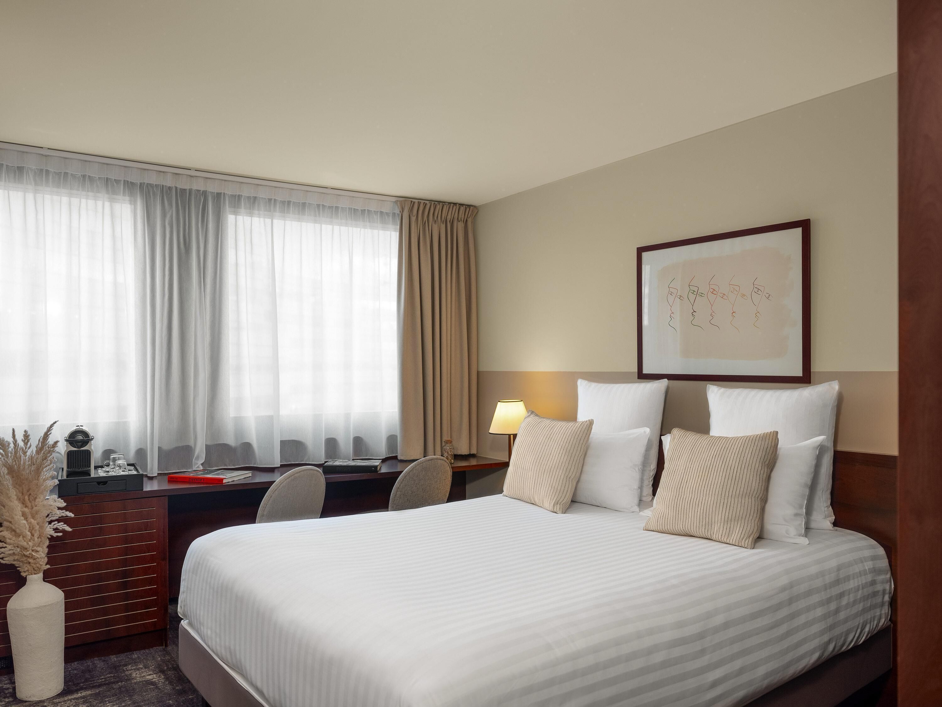 A hotel room with a bed, desk, chair, lamp, and artwork on the wall.