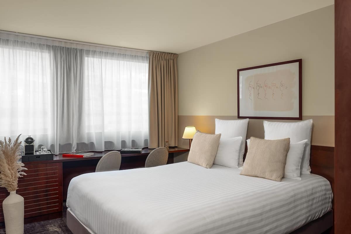 A hotel room with a bed, desk, chair, lamp, and artwork on the wall.