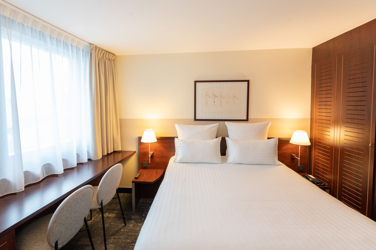 Habitación de hotel con cama, dos mesitas de noche con lámparas, un escritorio con silla, un ventanal con cortinas y un cuadro en la pared.