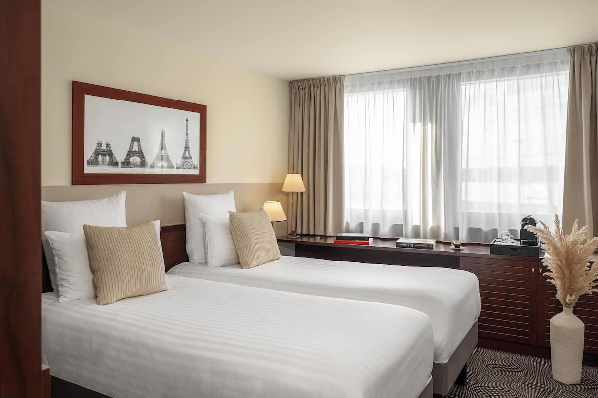 A hotel room with two beds, a framed picture of the Eiffel Tower, a nightstand with a lamp, and a window with sheer curtains.