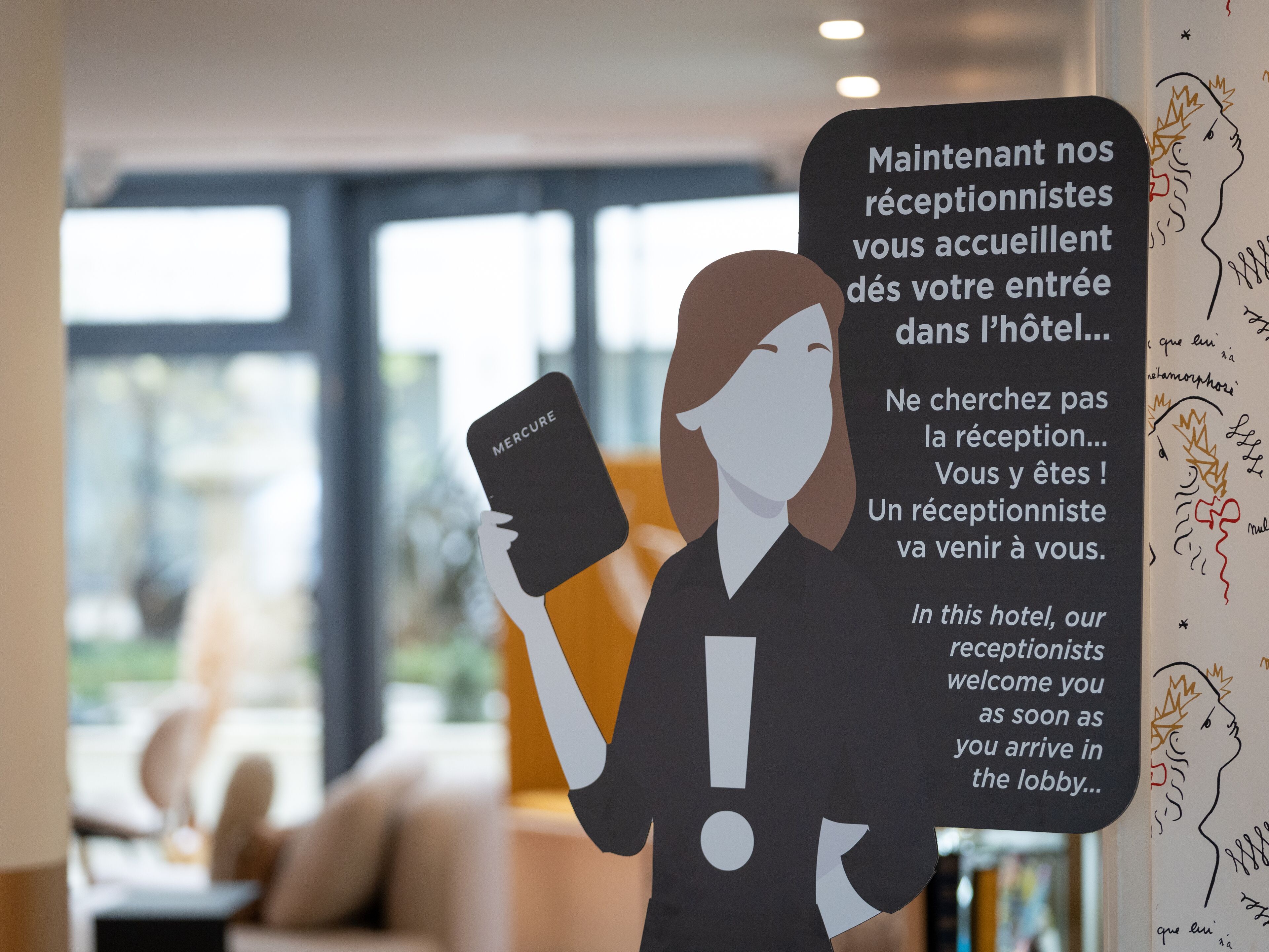 Una persona sosteniendo un cartel con 'MERCURE', de pie en el vestíbulo de un hotel con un gran ventanal y un sofá al fondo.
