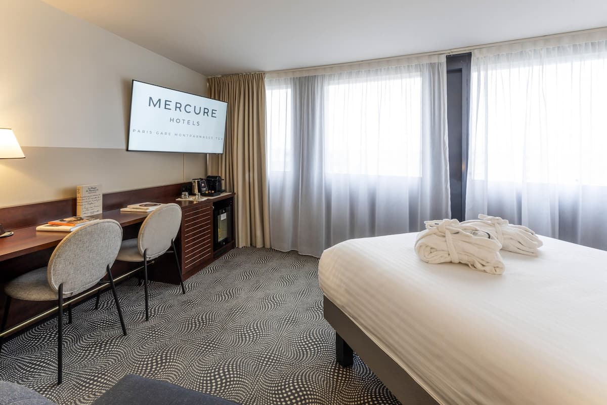A hotel room with a bed, a desk with a chair, a television displaying 'MERCURE HOTELS,' and a window with curtains.
