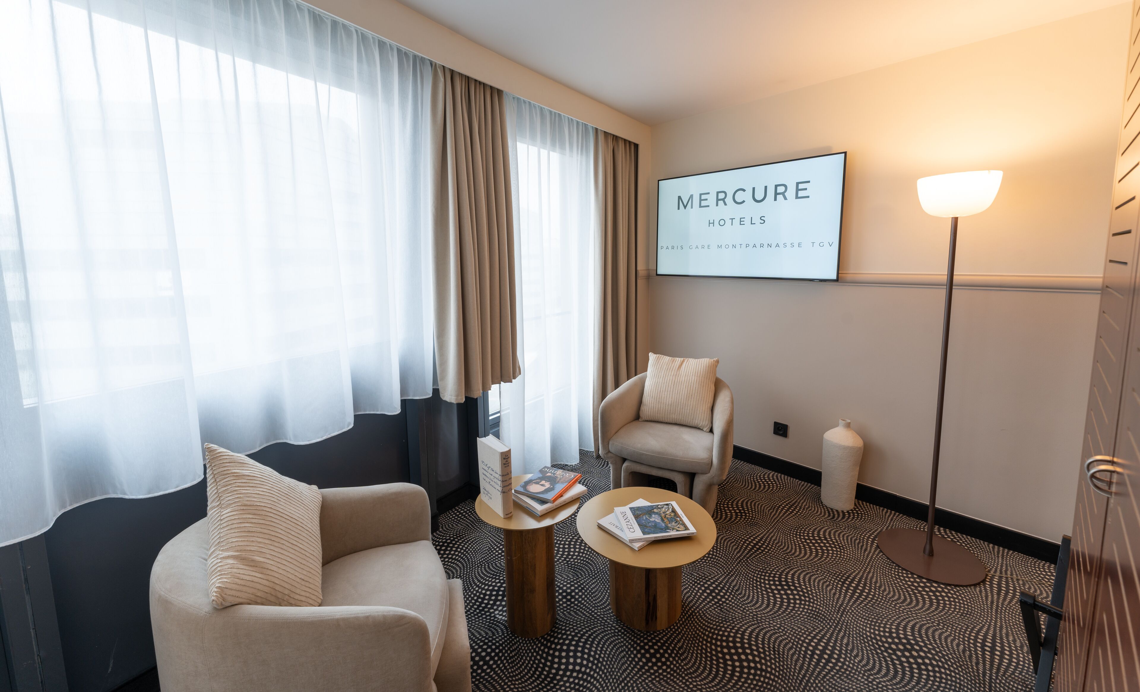 A hotel room with a sofa, armchair, two small tables, a lamp, and a sign reading "Mercure Hotels Paris Gare Montparnasse TGV".