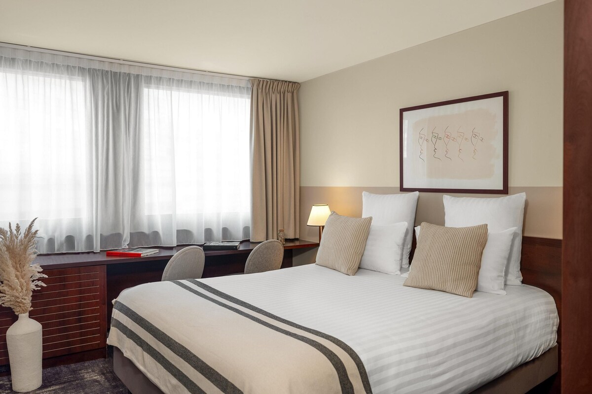 A hotel room with a bed, desk, chair, lamp, and a framed picture on the wall.