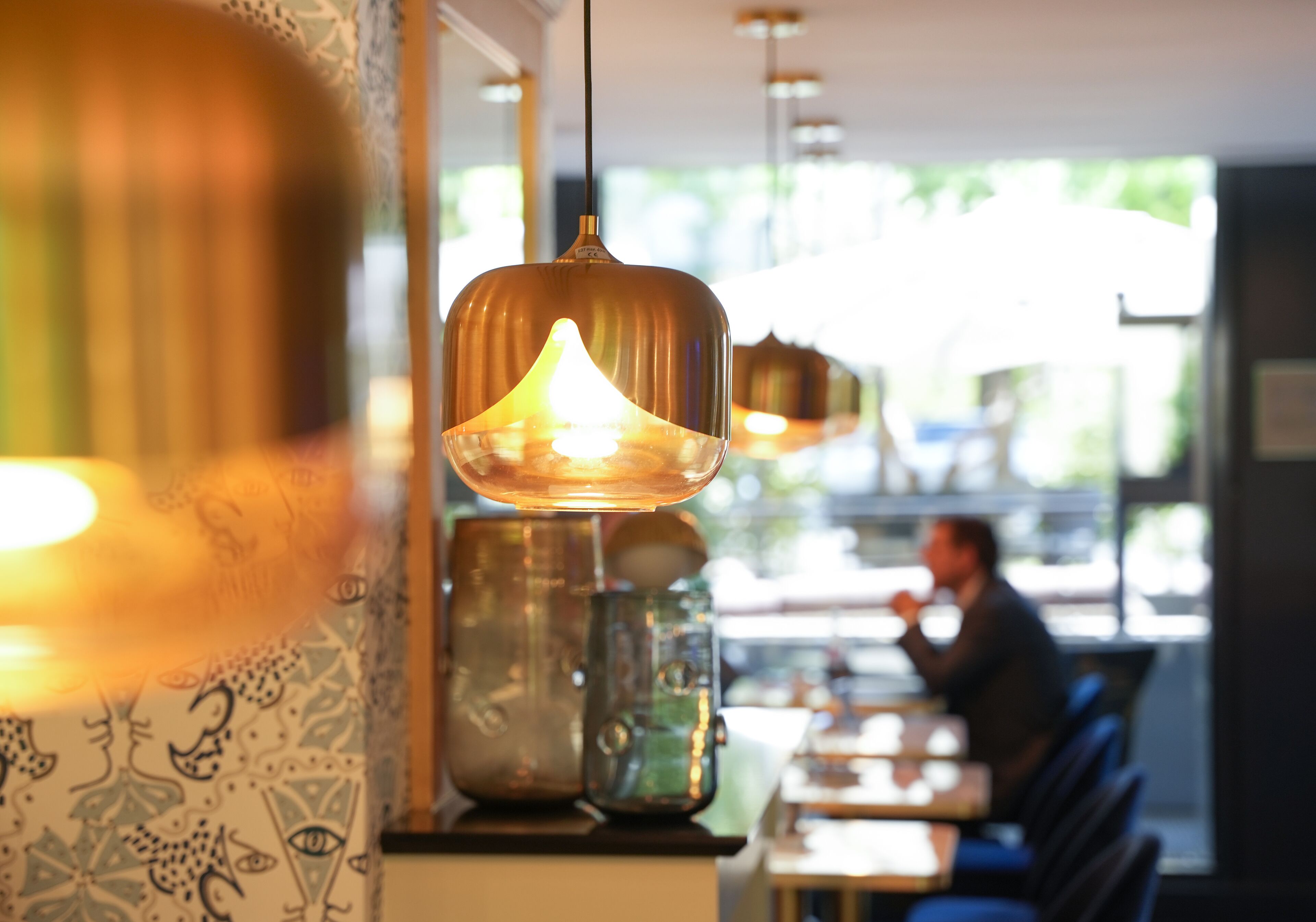 A restaurant interior with hanging pendant lights, patterned wallpaper, and a person dining in the background.