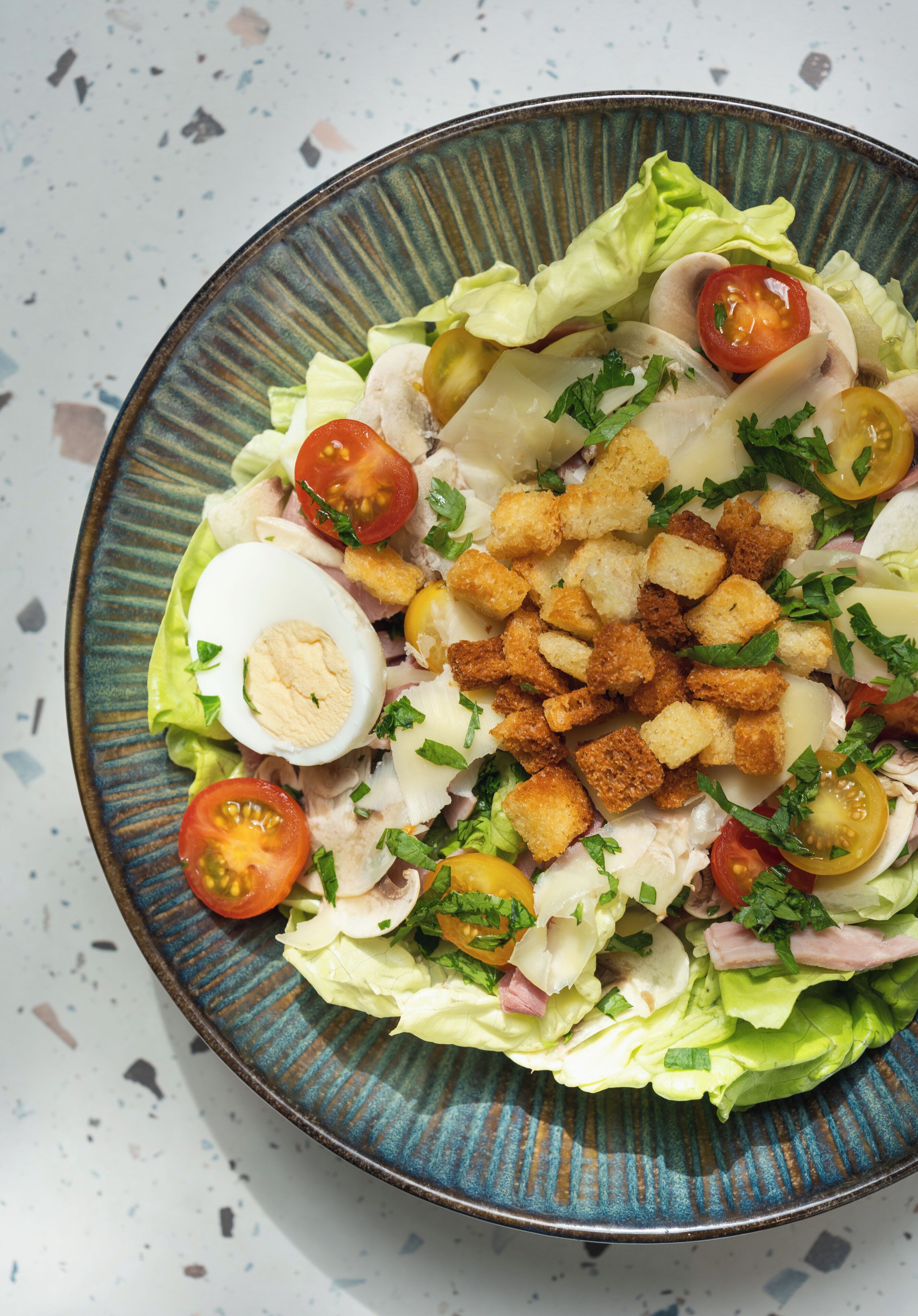 Een salade met sla, tomaten, een gekookt ei, croutons en kaas op een keramieken bord.