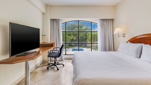 A hotel room with a bed, a desk with a television, a chair, and a large window offering a view of greenery and a pool. - Fiesta Inn Colima (Colima)