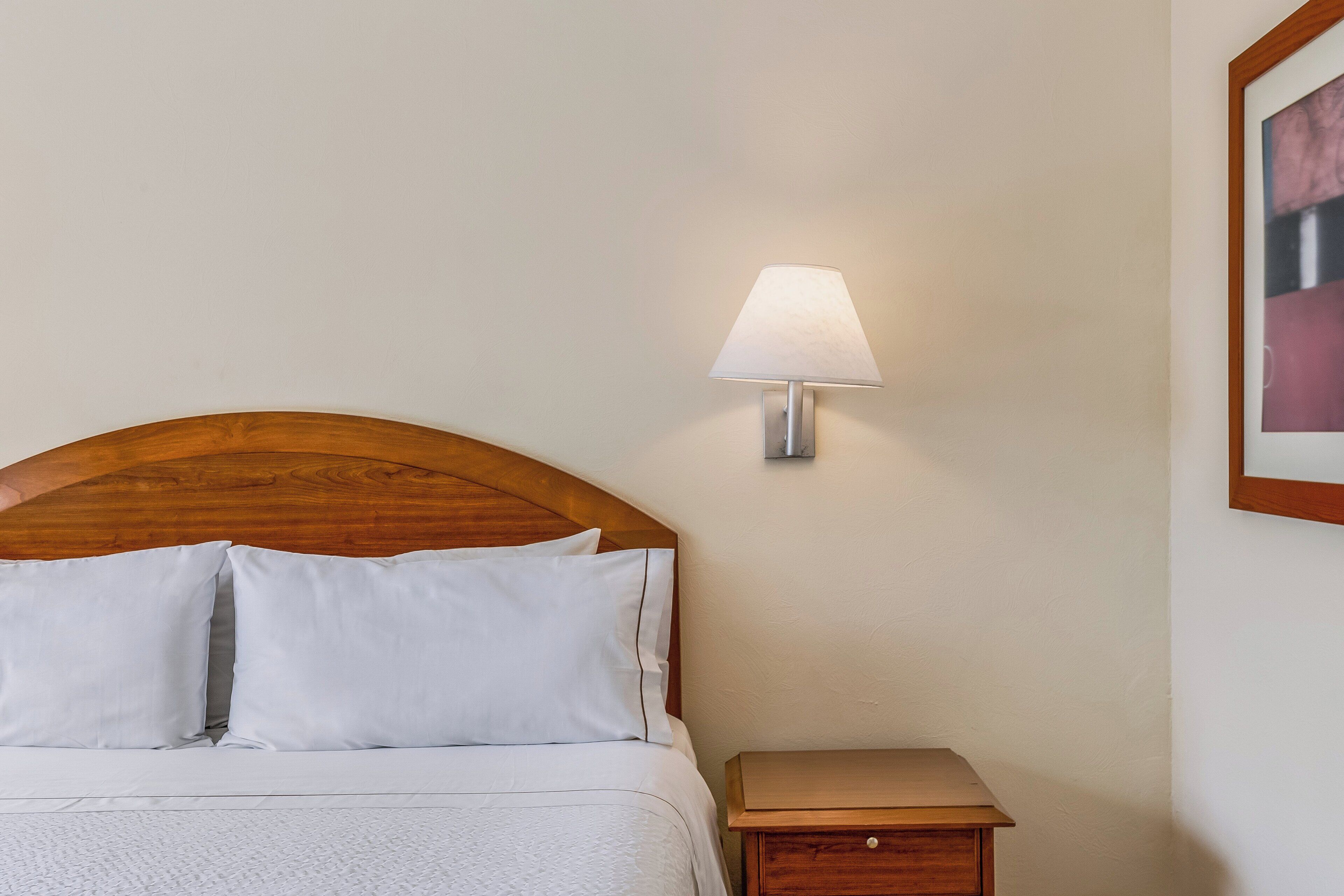A neatly made bed with white linens, a wooden headboard, a bedside table with a lamp, and a framed picture on the wall.