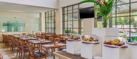 A spacious dining area with multiple tables and chairs, a buffet with various food items, and a large window offering a view of the outdoors.