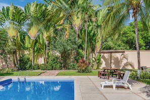 A swimming pool with lounge chairs and a picnic table, surrounded by lush greenery and palm trees.