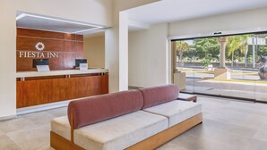 A modern hotel reception area with a wooden reception desk, a seating area with a red and white patterned bench, and a view of the street through large glass doors. - Fiesta Inn Colima (Colima)