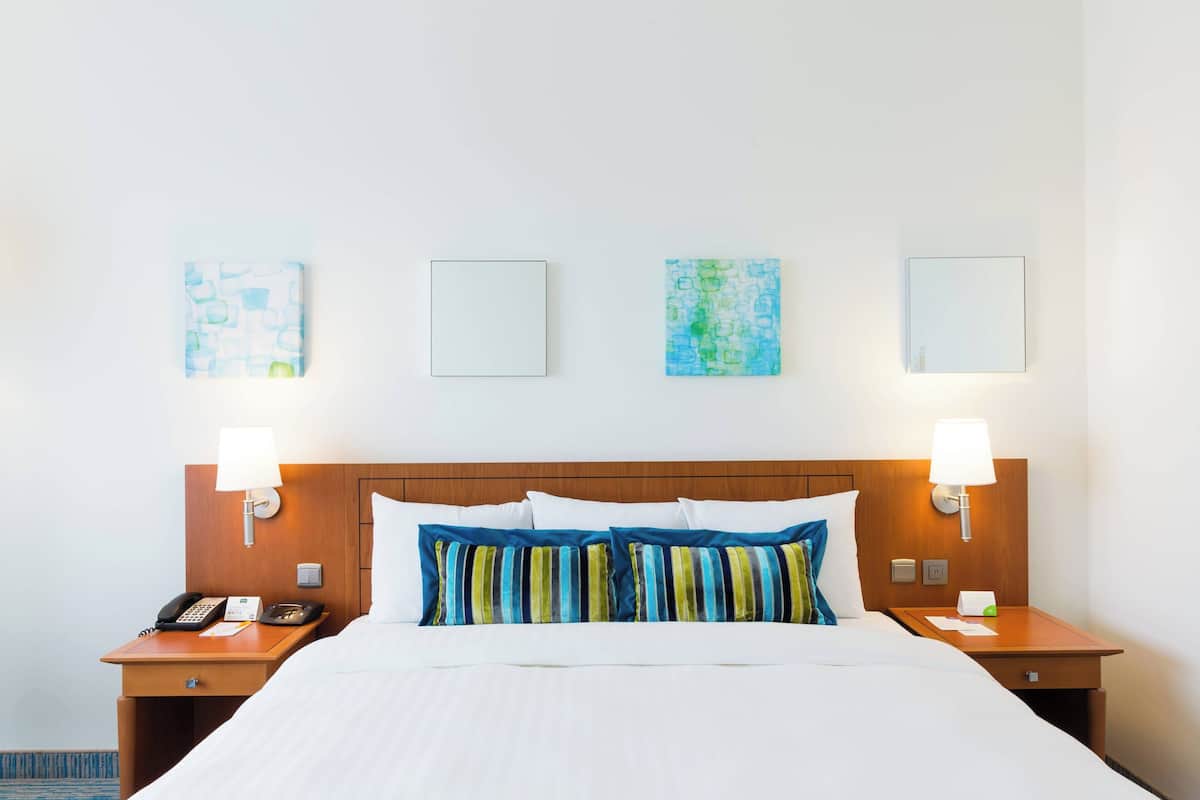 Ein Hotelzimmer mit Holz-Kopfteil, einem Bett mit weißer Bettwäsche, zwei Nachttischen mit Lampen und drei gerahmten Kunstwerken an der Wand.