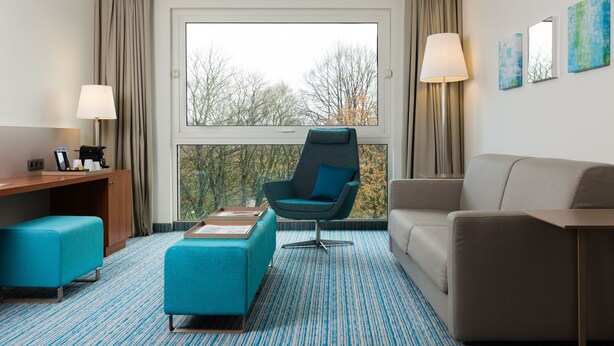 A hotel room with a large window, a desk with a lamp, a blue armchair, a sofa, and a small ottoman. - Courtyard by Marriott Brussels (Brussels)