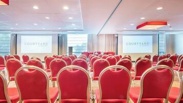 Un salón de conferencias con filas de sillas rojas, una pantalla grande mostrando 'COURTYARD BY MARRIOTT' y vistas de la ciudad a través de amplias ventanas.