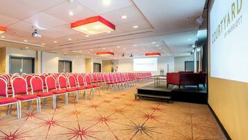 Une salle de conférence avec des rangées de chaises rouges, un tapis à motifs géométriques et une scène également dotée de chaises rouges.