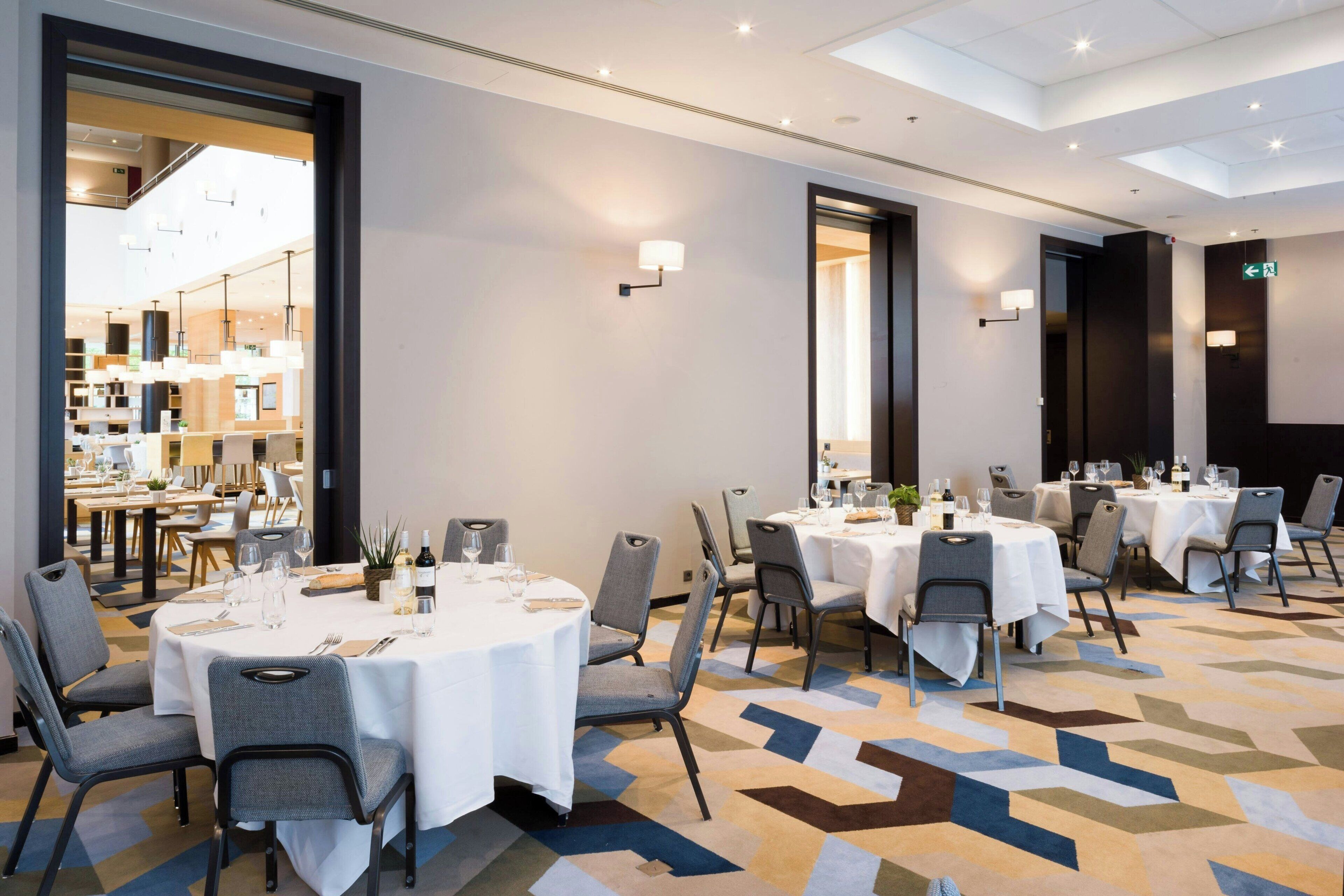 A hotel dining area with round tables, chairs, and a patterned carpet.