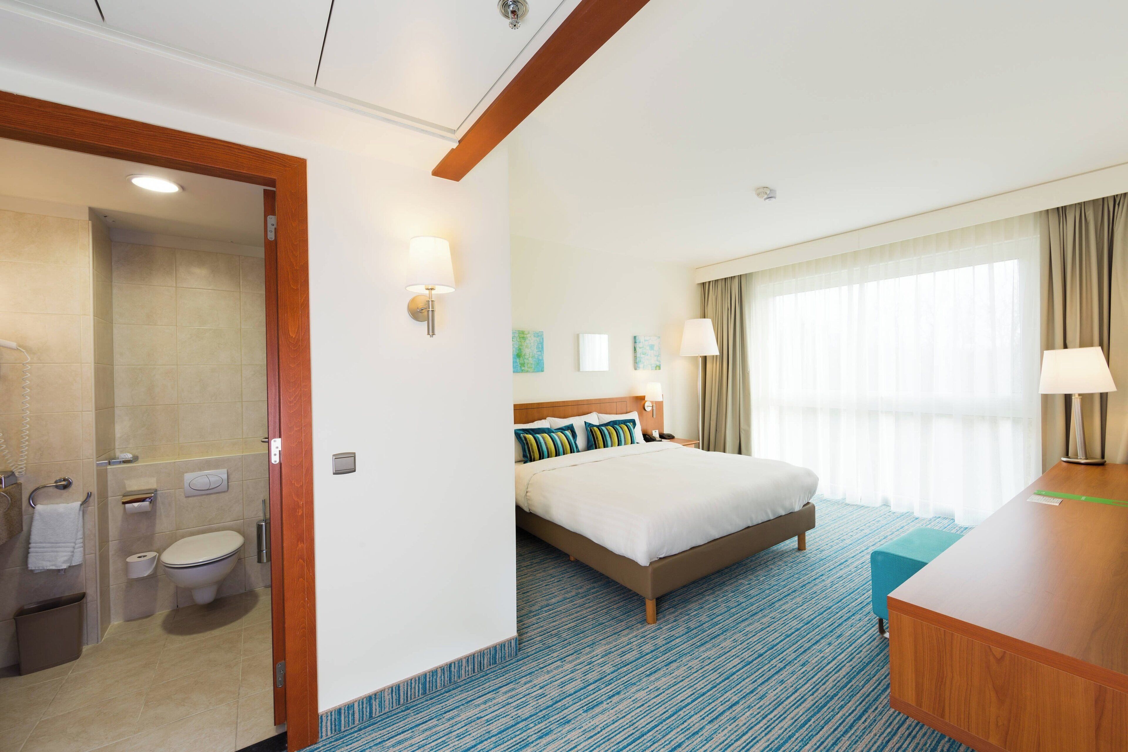 A hotel room with a bed, a desk, a bathroom, and a large window.
