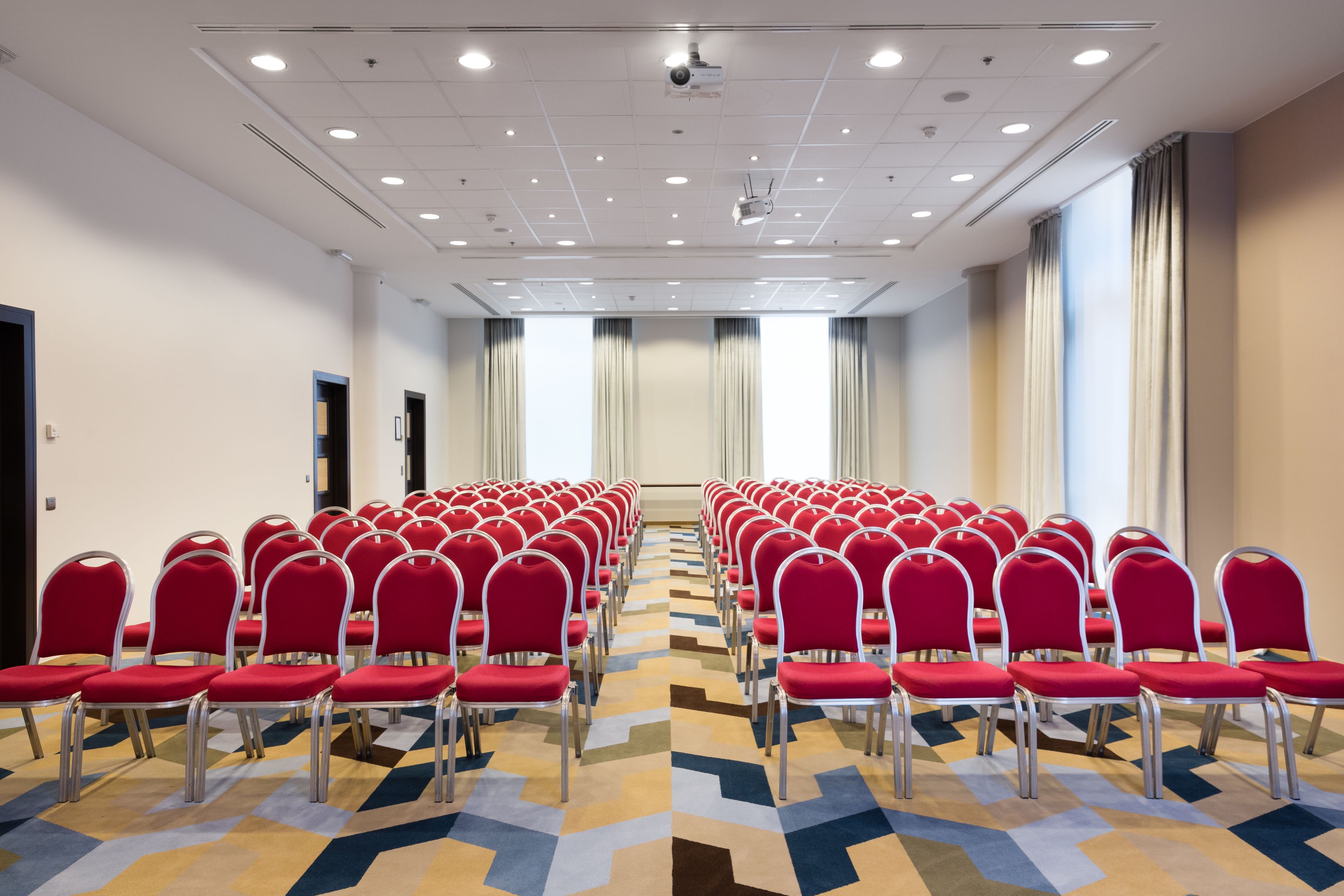 Un salón de conferencias con filas de sillas rojas, una alfombra estampada y ventanas grandes con cortinas.