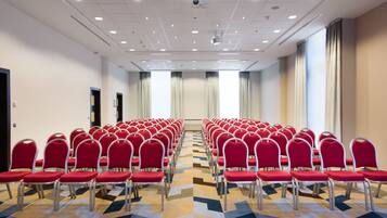 Un salón de conferencias con filas de sillas rojas, una alfombra estampada y ventanas grandes con cortinas.