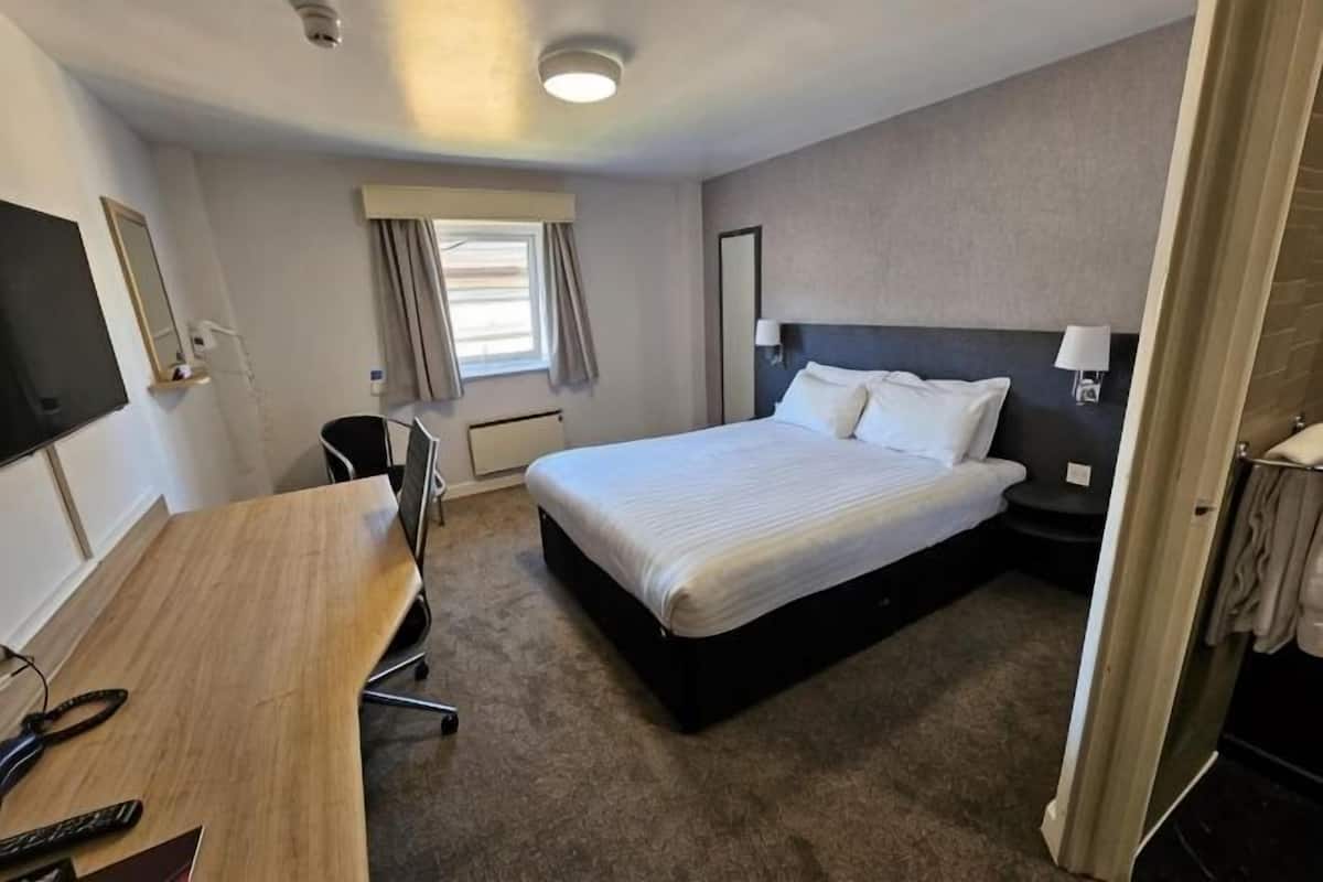 Quarto de hotel com cama, escrivaninha, cadeira, televisão e janela com cortinas.