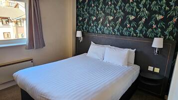 A hotel room with a large bed, two bedside lamps, a window with curtains, and a wall with a leafy pattern.