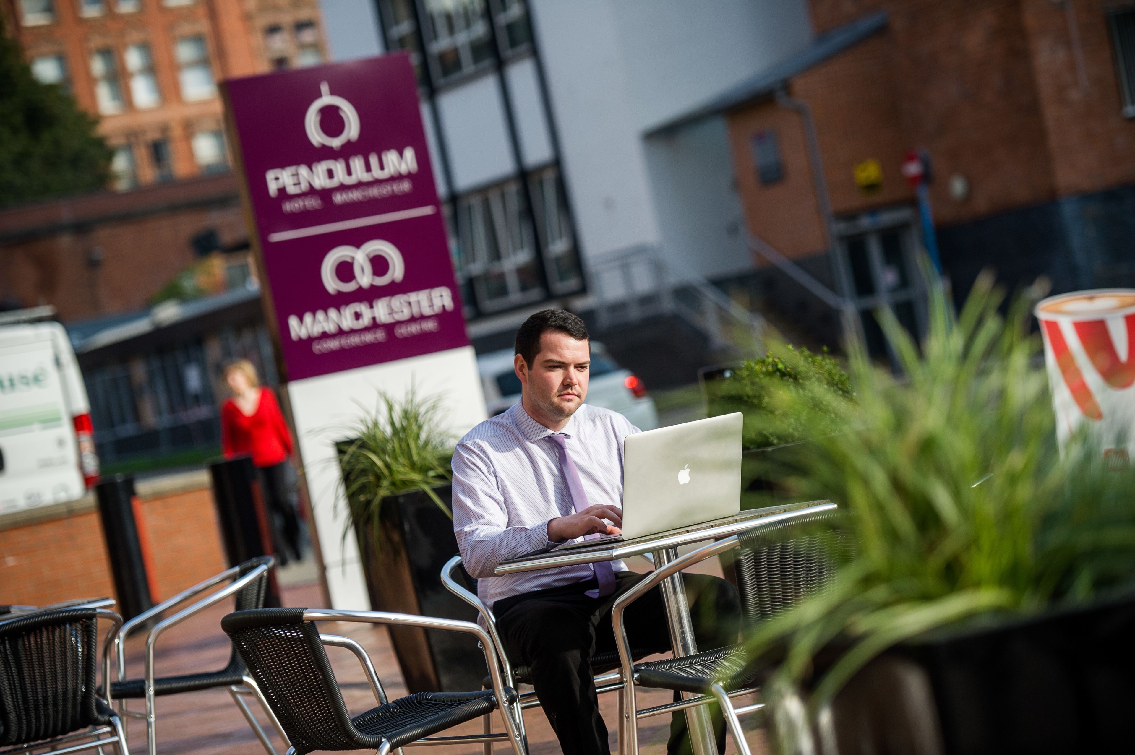 A man using a laptop at an outdoor table with chairs, in front of a sign for Pendulum Manchester.