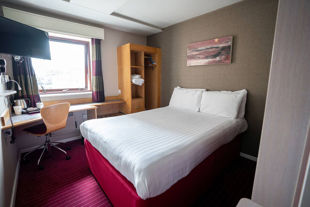 Quarto de hotel com cama grande, uma escrivaninha, uma cadeira, guarda-roupa e um quadro na parede.