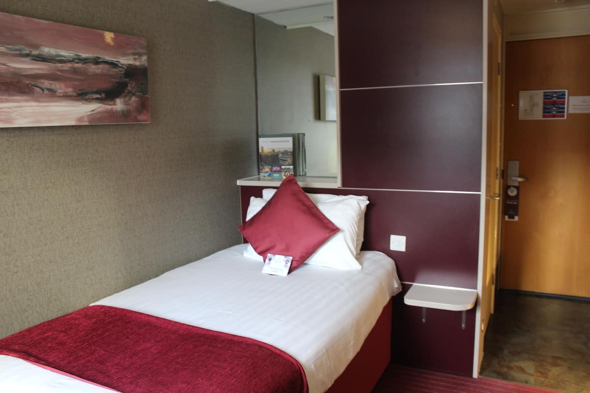 Quarto de hotel com cama, manta vermelha, travesseiro, mesa de cabeceira pequena, espelho e porta com um aviso.