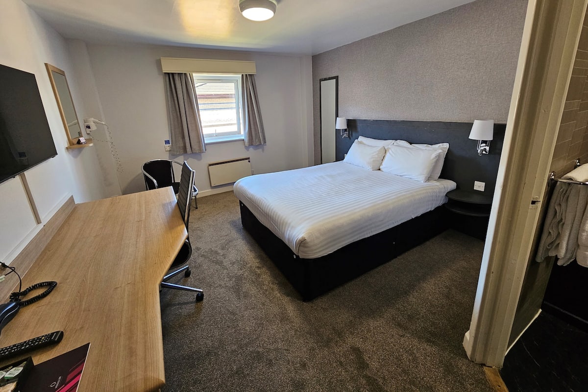 Quarto de hotel com cama, escrivaninha, cadeira, televisão, janela com cortinas e luminária na parede.