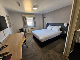 Habitación de hotel con cama, escritorio, silla, televisor, ventana con cortinas y lámpara de pared.