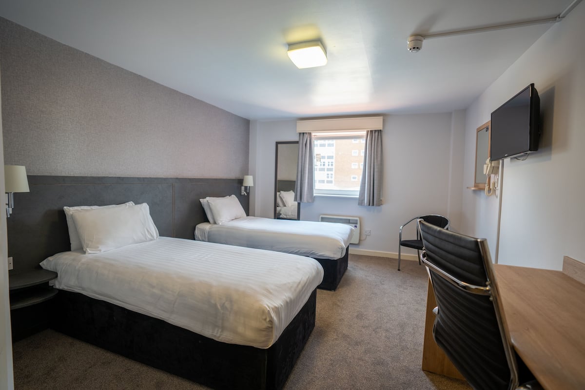 Quarto de hotel com duas camas, uma mesa, uma cadeira, televisão, janela com cortinas e luminária no teto.