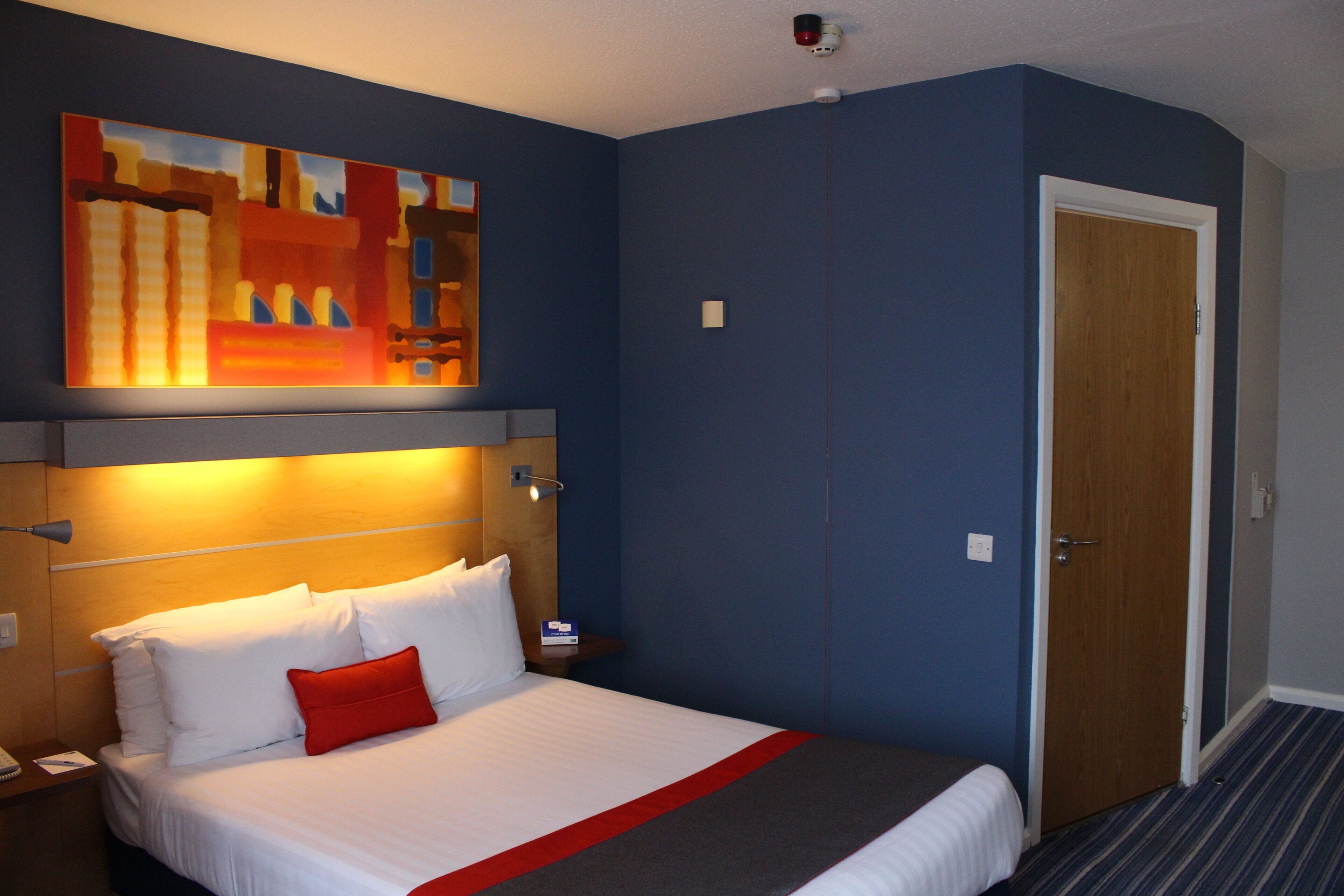 A hotel room with a bed, a nightstand, a wall-mounted lamp, a painting, and a door.