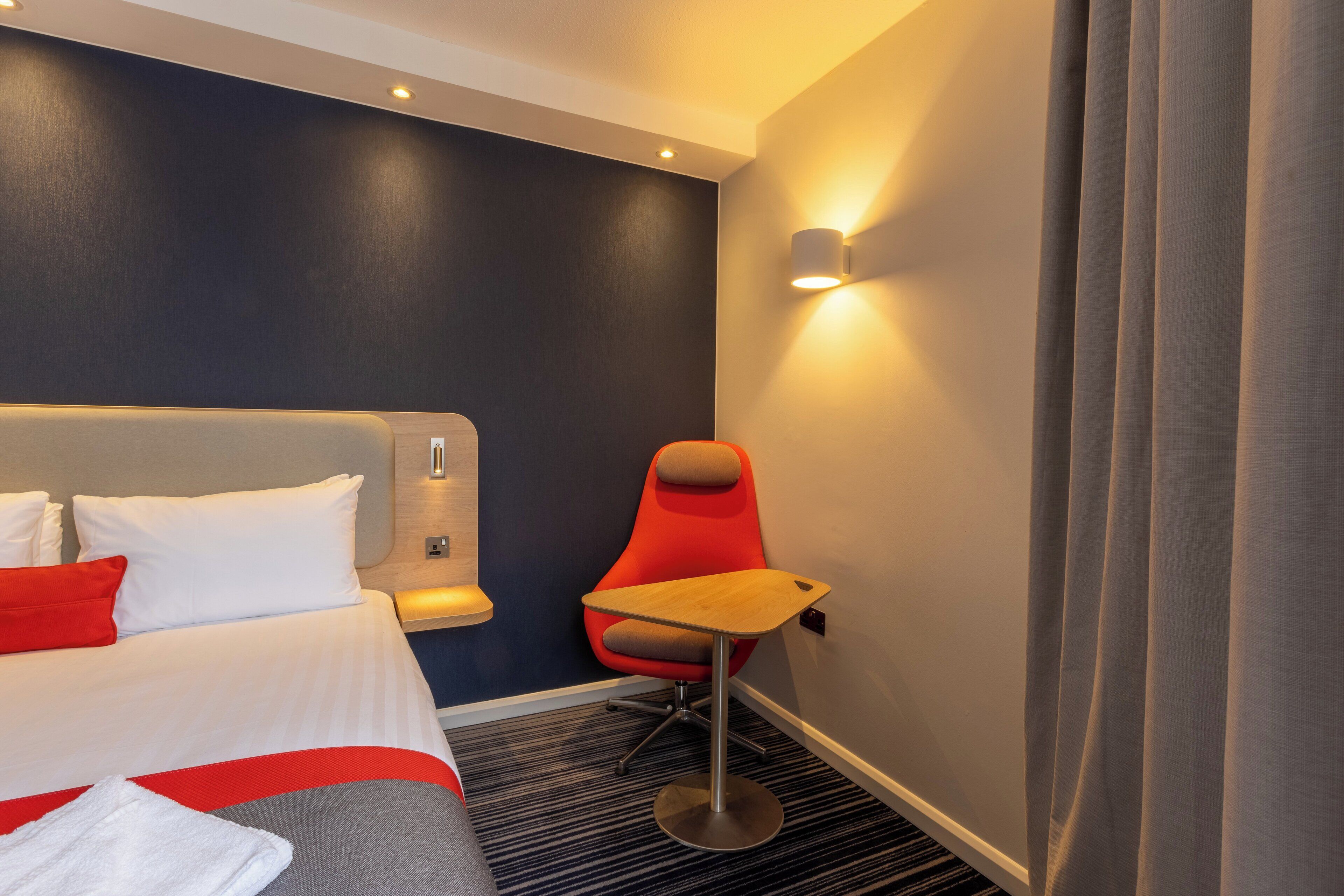 A hotel room with a bed, a red chair, a small table, and a wall-mounted light.