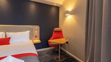 A hotel room with a bed, a red chair, a small table, and a wall-mounted light.