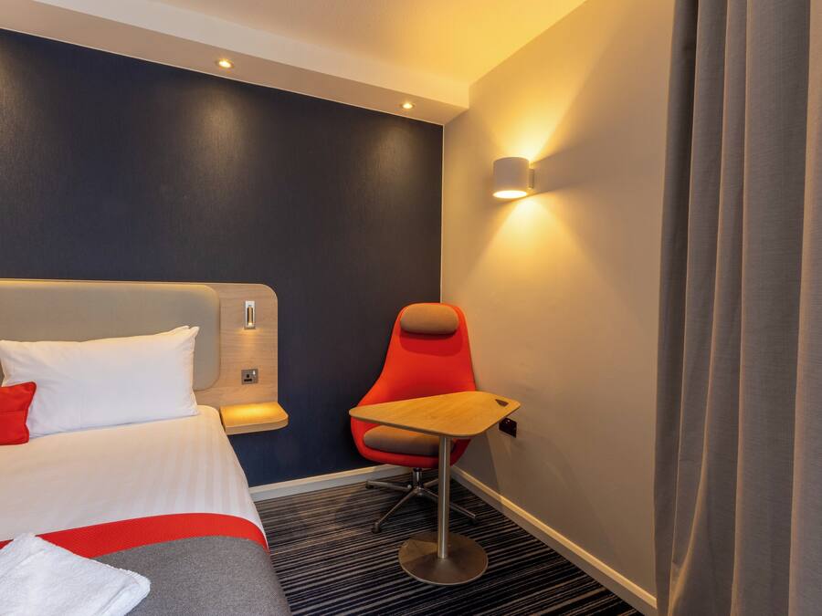 A hotel room with a bed, a red chair, a small table, and a wall-mounted light.