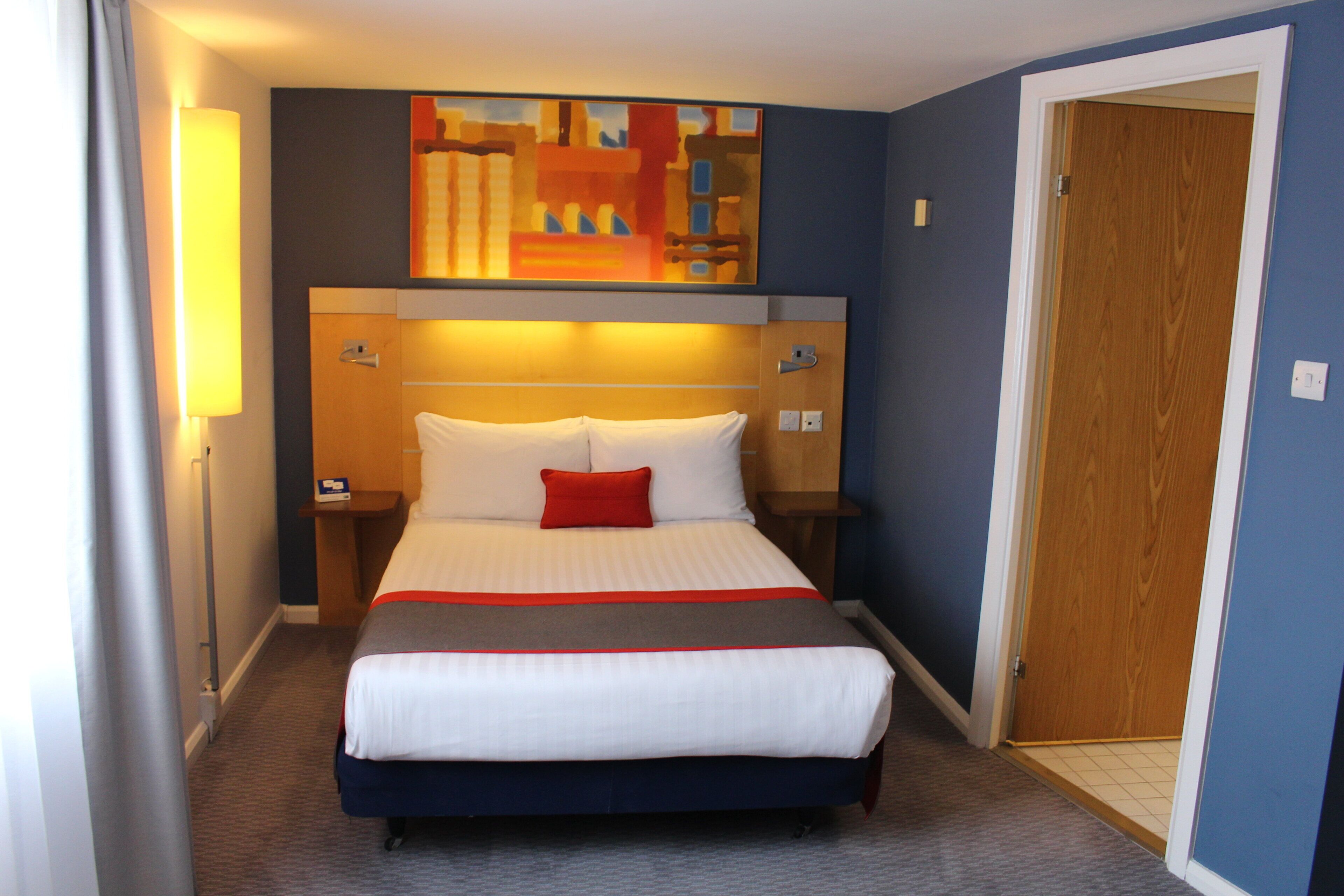 A hotel room with a bed, nightstands, a painting above the bed, and a door.