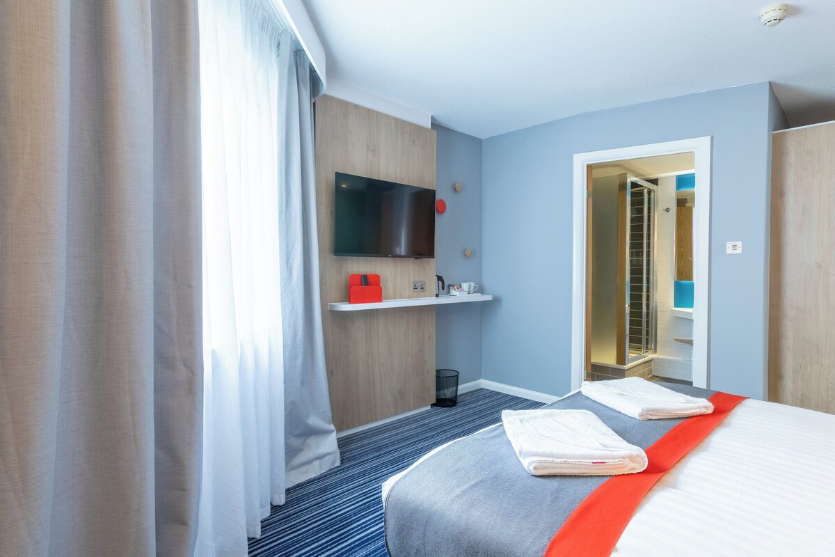 Une chambre d’hôtel comprenant un lit, un téléviseur fixé au mur, une petite étagère avec quelques objets, et une porte donnant accès à la salle de bain.