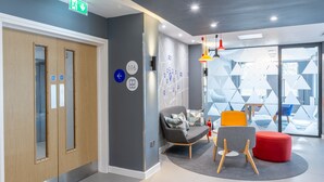 A modern lobby with a wooden door, a grey sofa, a red ottoman, and a glass wall with geometric patterns.
