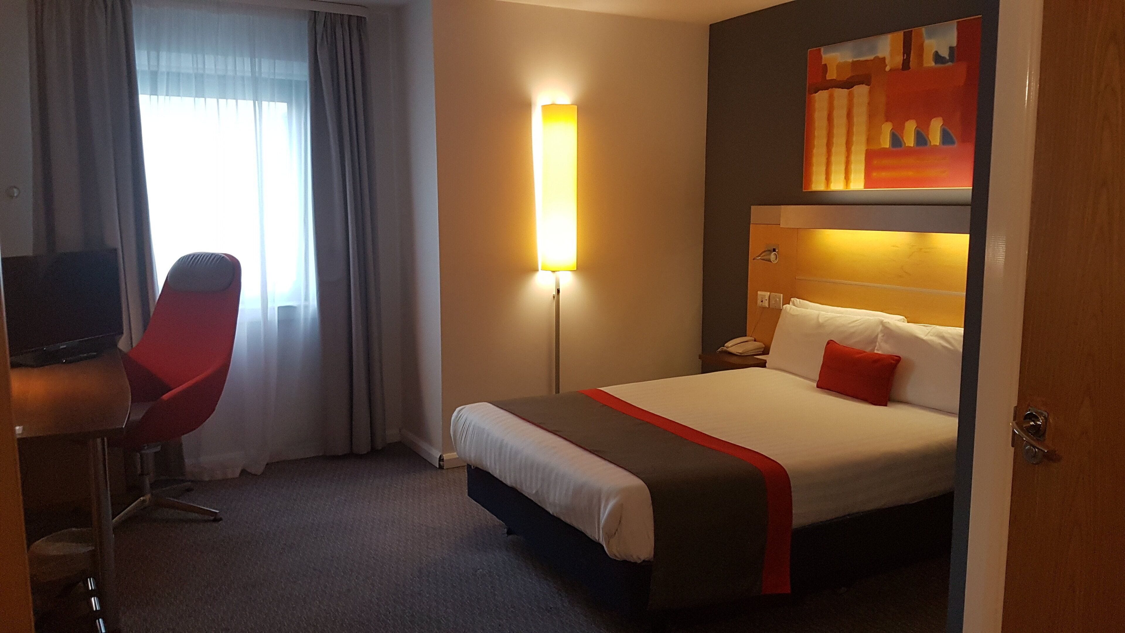 Una habitación de hotel con una cama, un escritorio con una silla, una mesita de noche con un teléfono y un cuadro en la pared.