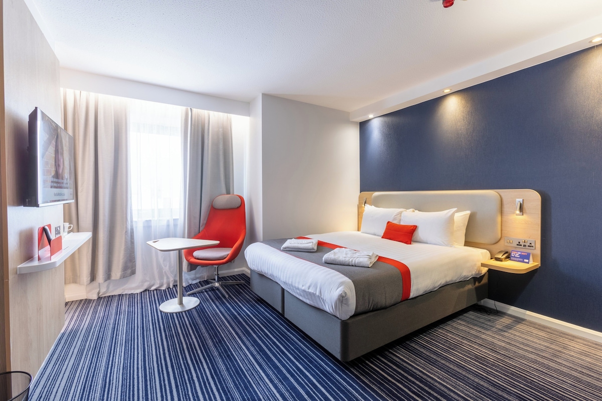 A hotel room with a bed, a desk, a chair, a TV, and a window with curtains.