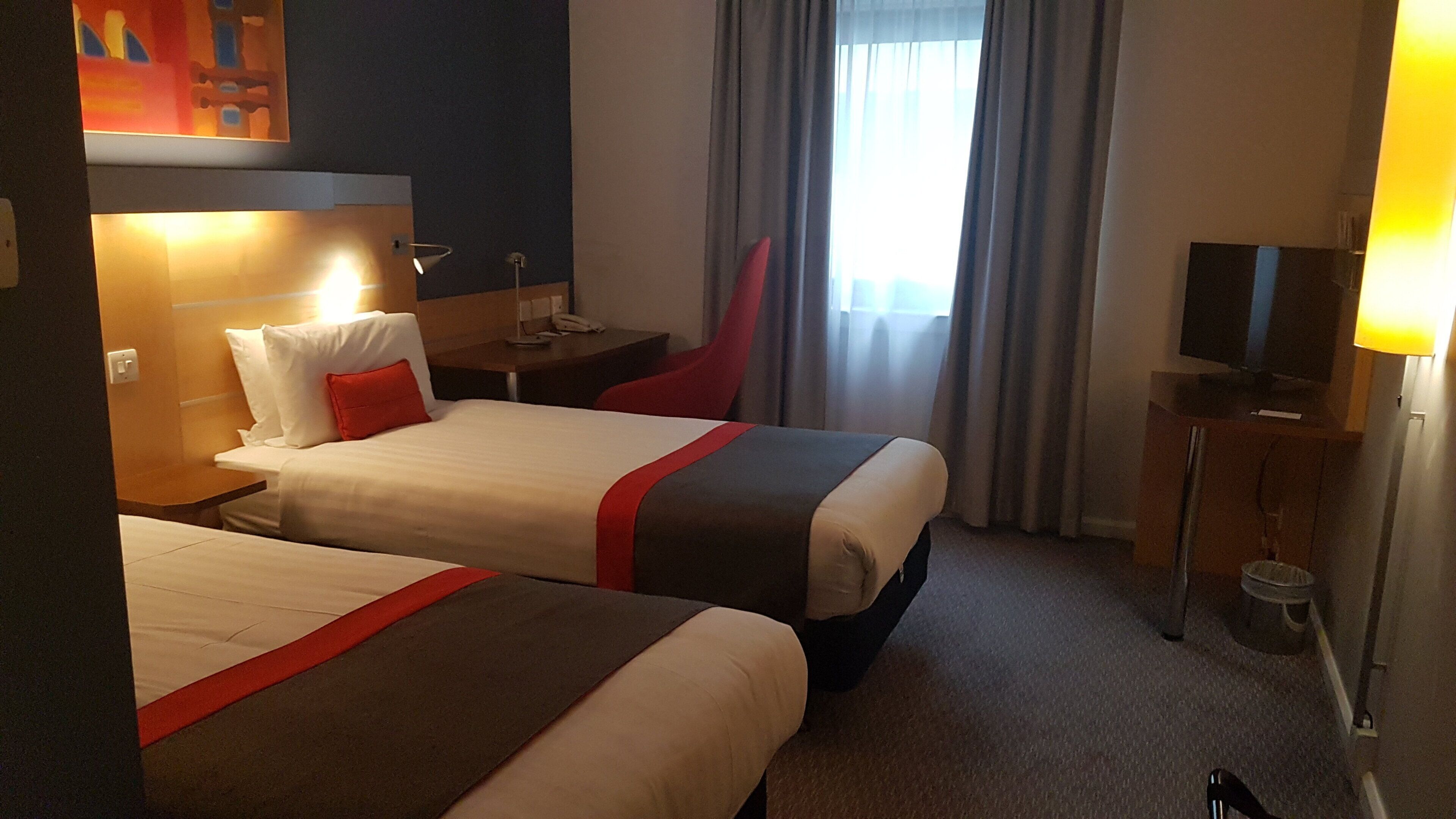 Ein Hotelzimmer mit zwei Betten, einem Schreibtisch, einem Stuhl, einem Fernseher und einem Fenster mit Vorhängen.