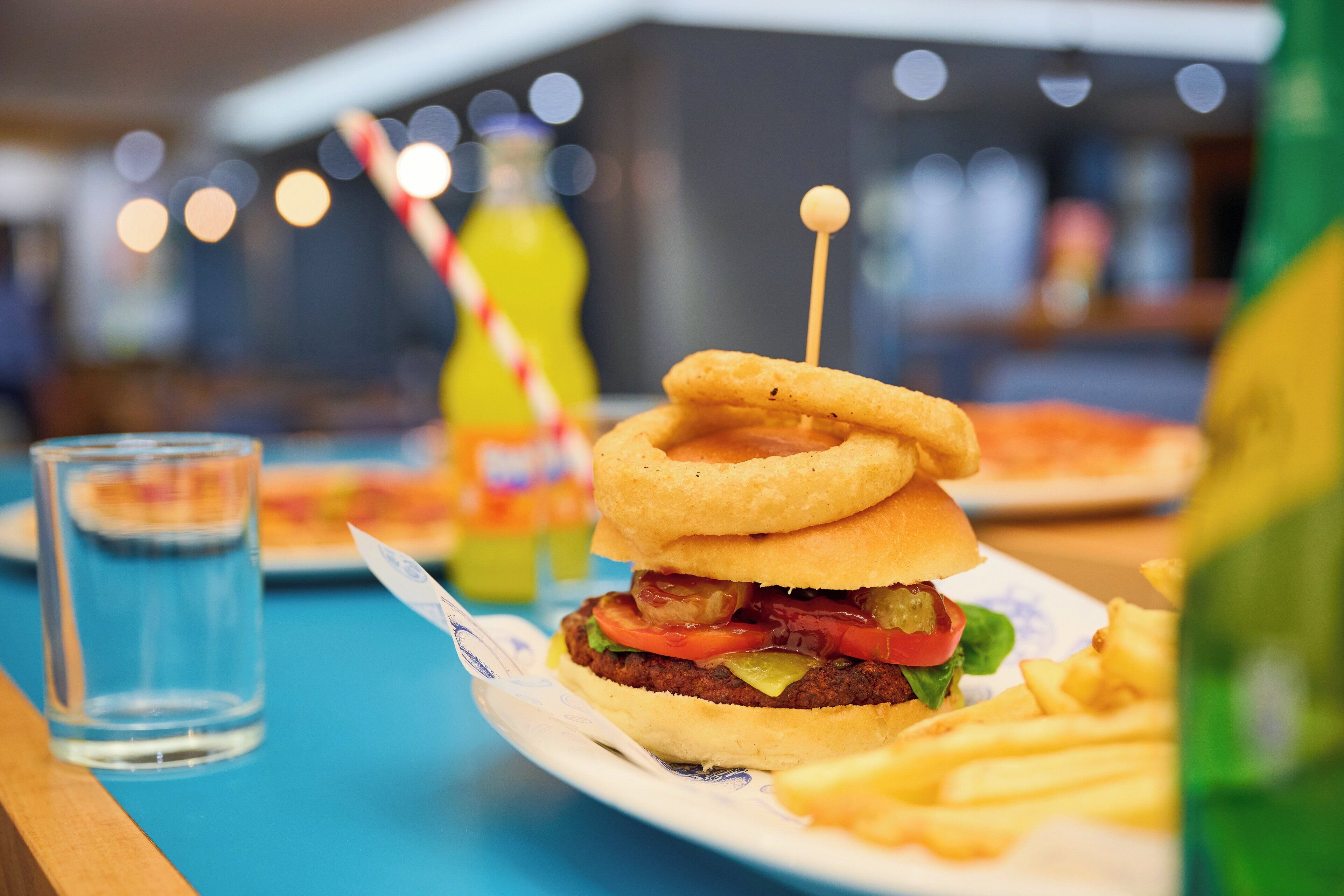 A burger with a toothpick, fries, a drink, and a bottle on a table.