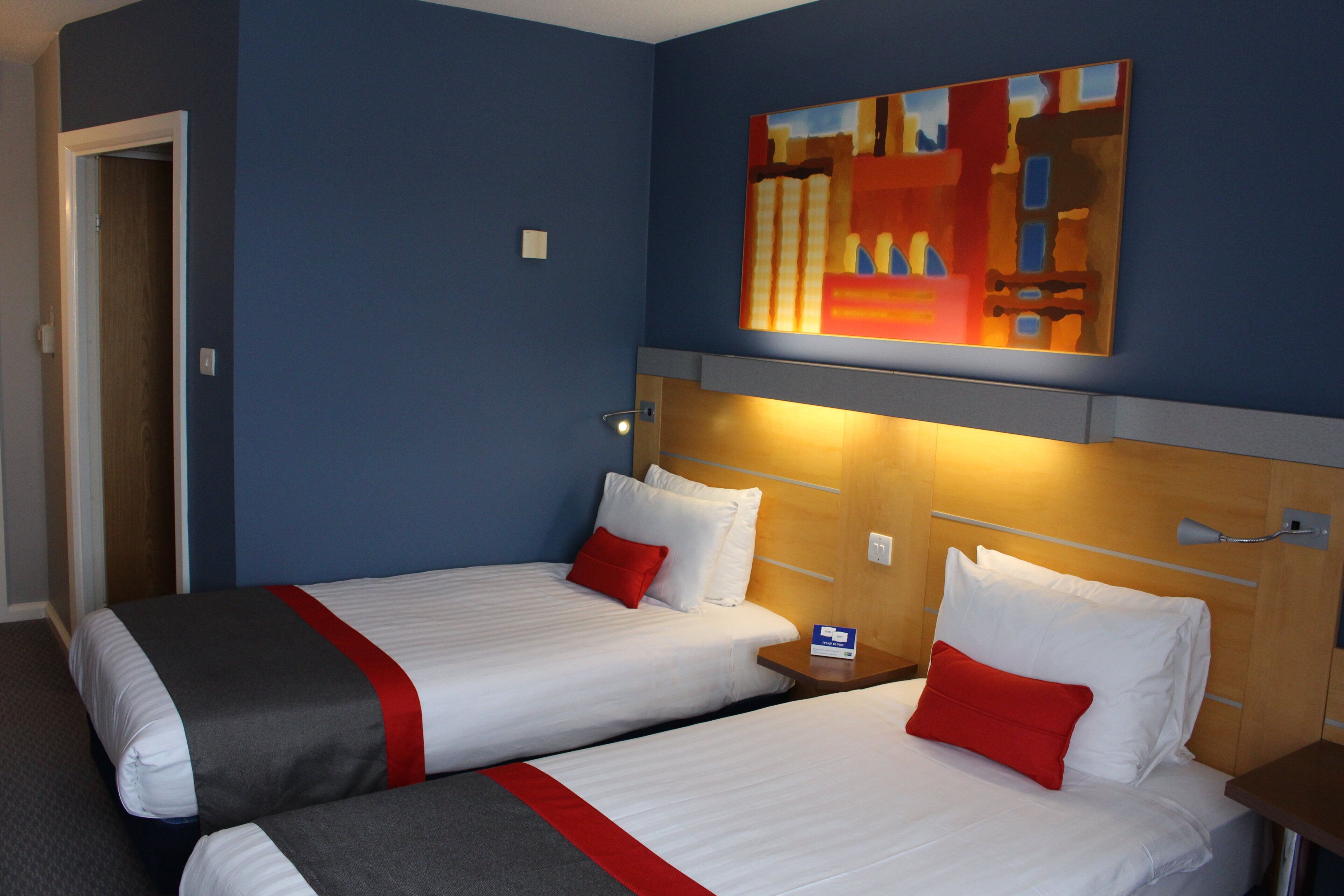 A hotel room with two beds, each with white bedding and red accents, a wooden headboard, and a blue wall.
