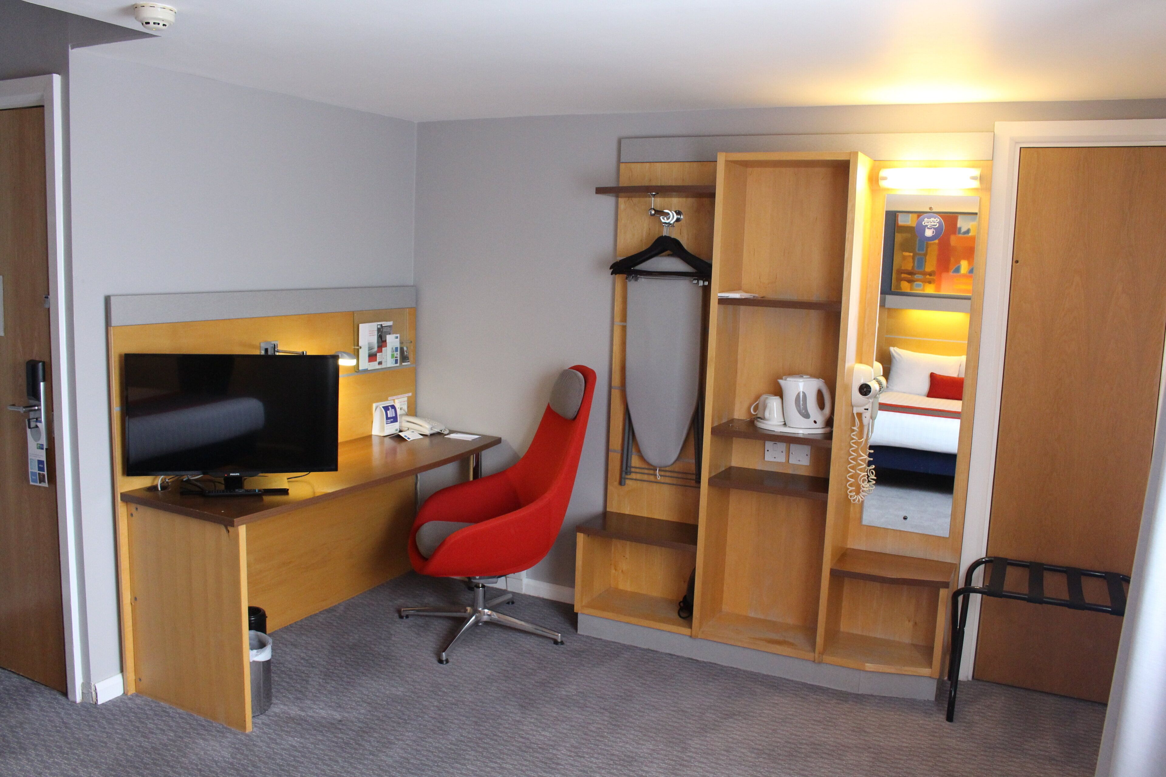 A hotel room with a television, a red chair, a desk, a bed with a headboard, and a wardrobe.
