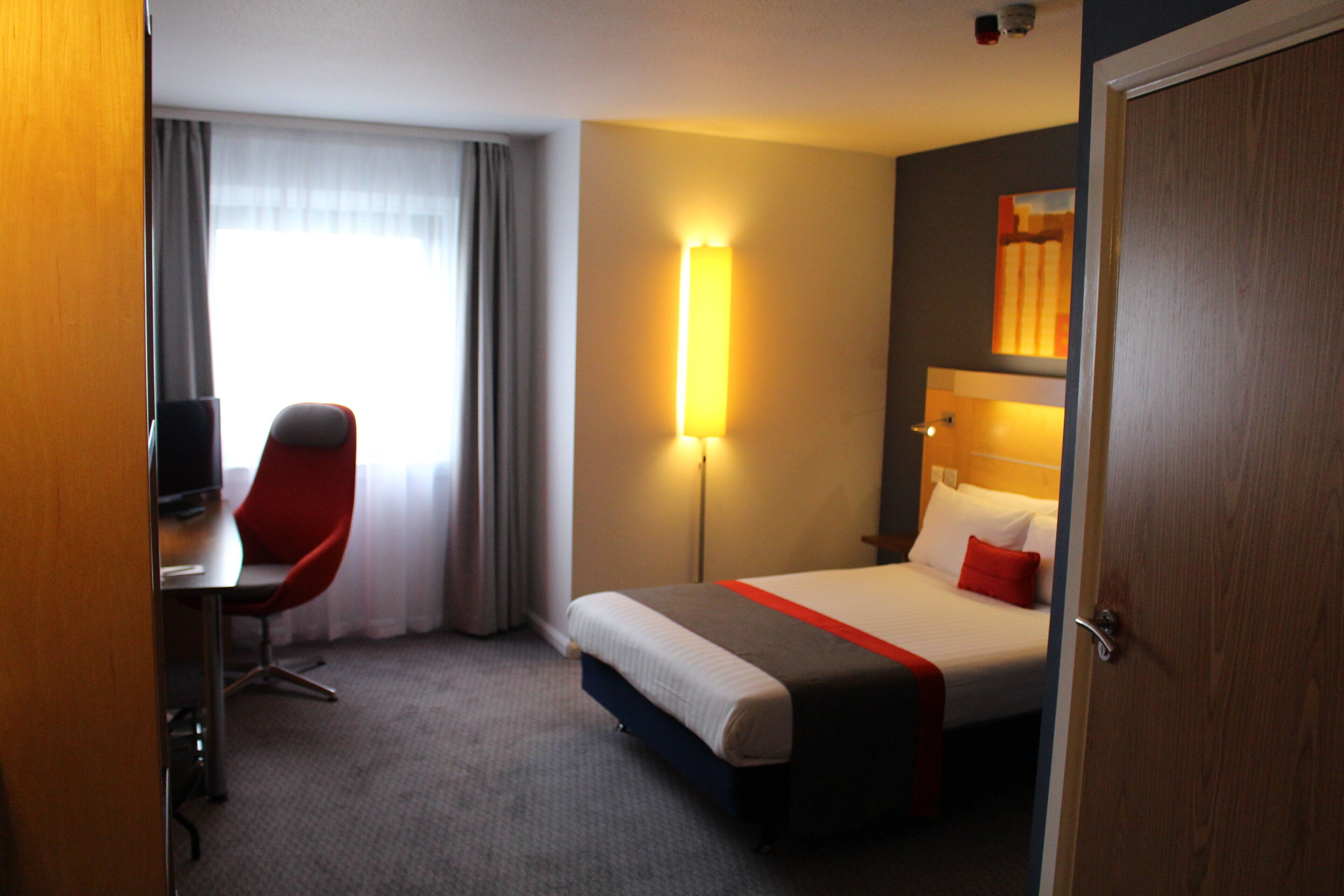 A hotel room with a bed, a desk, a chair, and a bedside table.