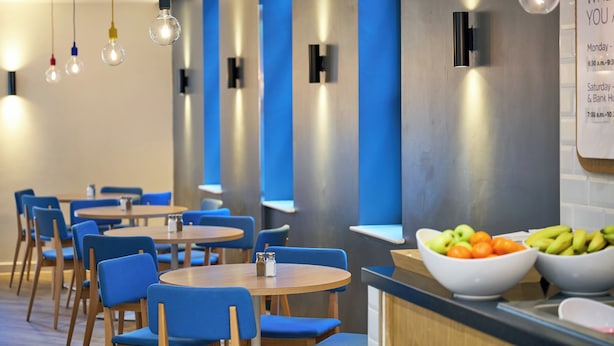 A modern restaurant with blue chairs and tables, hanging light bulbs, and a fruit bowl on the counter.