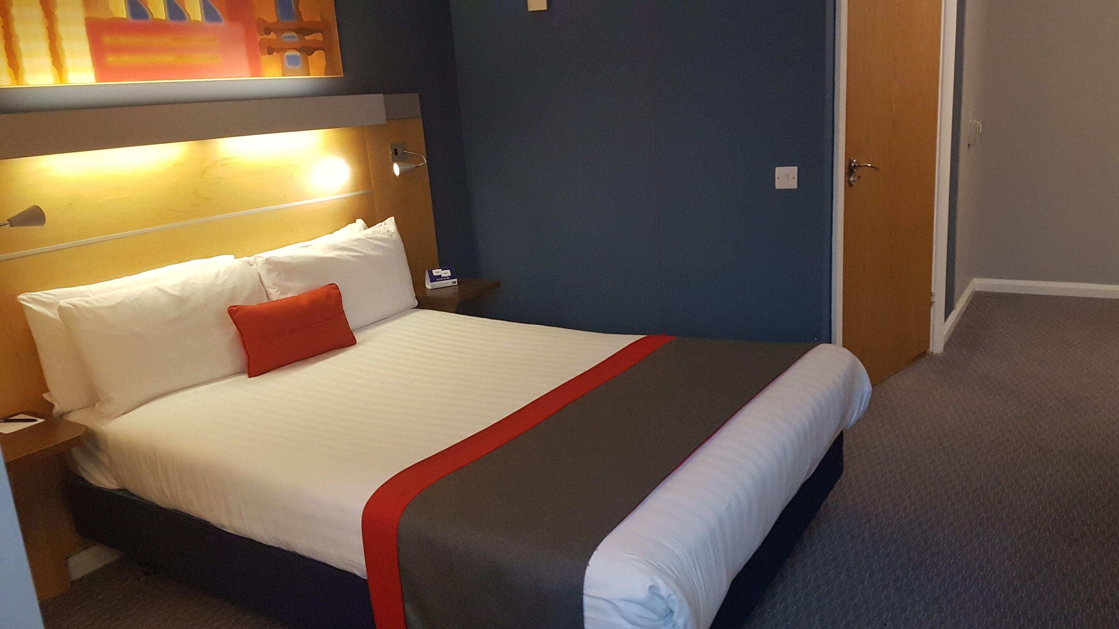 Una habitación de hotel con una cama, una mesita de noche, una lámpara y una puerta.