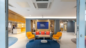 A modern lobby with a reception desk, a blue wall with 'Always Welcome...' text, a sofa, and two yellow chairs.