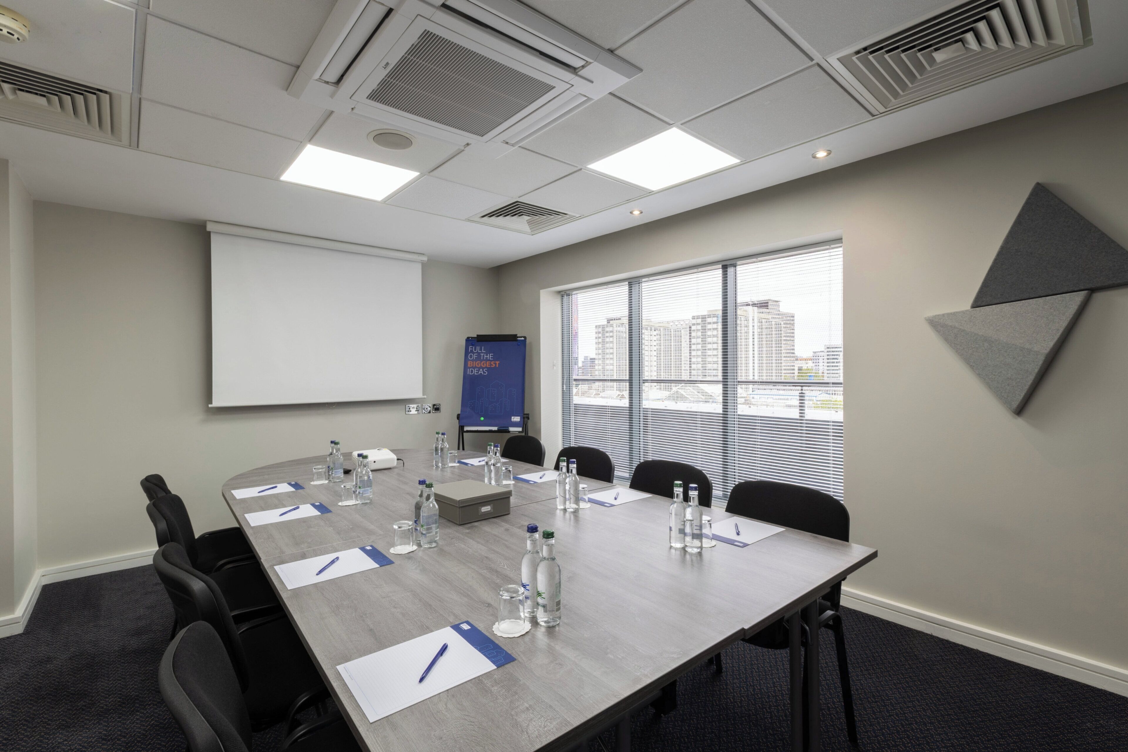 A conference room with a long table, chairs, a projector screen, and a window with blinds.